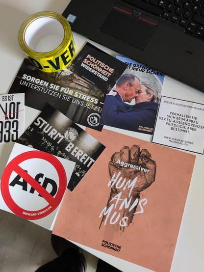 Eine Auswahl an Stickern. Man sieht schlichte Sticker wie AfD durchgestrichen oder den Text "Verhalten Sie sich beim Abbau von EU-Außengrenzen friedlich, aber bestimmt". Dazu auch aufwändigere, wie z. B. ein KI generiertes Bild, wie sich Friedrich Merz und Alice Weidel in den Armen liegen. Darüber steht "Die Grenze ist nicht mehr sicher". Außerdem ist eine große Karte mit einer nach oben gestreckten Faust zu sehen, die mit "Agressiver Humanismus" beschriftet ist. Oben links im Bild liegt die erwähnte Rolle Klebeband, wo der Text "AfD-Verbot.de" zu erahnen ist.
