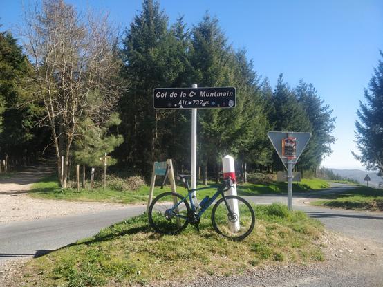 Photo de mon vélo bleu posé sur la pancarte du col de la croix de Montmain, avec des sapins derrière. Le ciel est bleu sans aucun nuages.