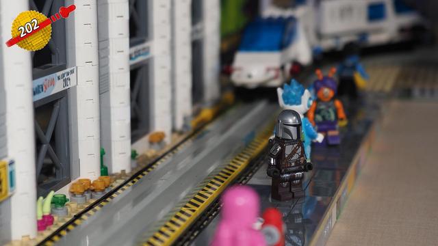 Man sieht einen aus Klemmbausteinen gebauten Weg, welcher neben einer Schiene verläuft. Auf dem Weg stehen mehrere Minifiguren und im Hintergrund sieht man einen Zug.