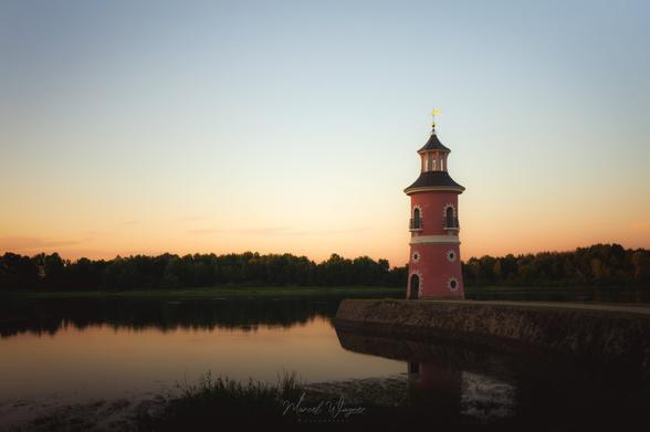 A red, round tower with a dark roof stands on a stone pier extending into a calm lake, with the water reflecting the tower and a pastel sunset sky. Trees form a dark green backdrop along the horizon.