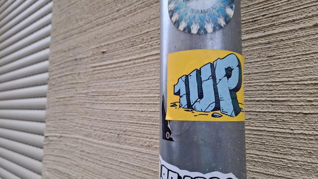 Graues Regenrohr vor einer beigen Wand. Mehrere Aufkleber, in der Mitte ein gelber Aufkleber nit blauem "1 Up" in Steinoptik.