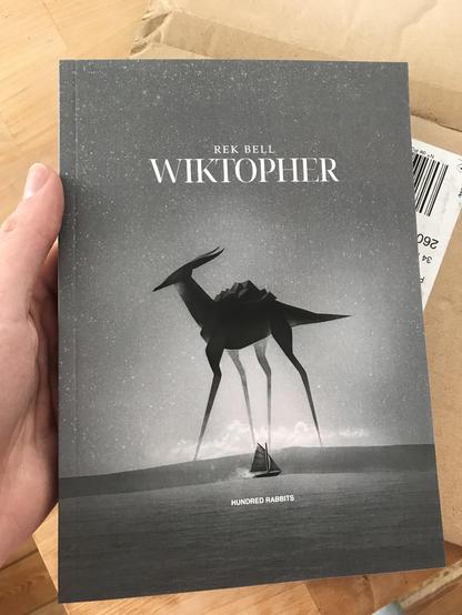 Photograph of the novel Wiktopher by Rek Bell, held my hand. The cover depicts a tiny sailboat on the sea, in grey weather. In the background is a giant black creature.