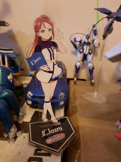 An acrylic stand of Riko Sakurauchi in a Saitama Lions uniform inspired idol outfit. She is holding a dark blue flag that says Lions on it. Behind her is a Tamiya model of the Subaru Impreza WRX STi.