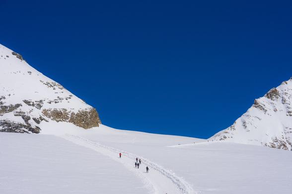 The image shows a snowy landscape with a clear, deep blue sky. There are people walking along a path in the snow between two mountain peaks. The terrain is predominantly covered in snow, with rocky outcroppings visible on the mountains. The scene suggests a peaceful winter hiking or mountaineering excursion.