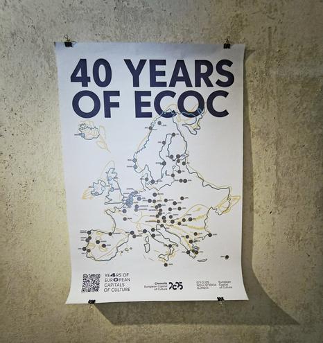 Poster an einer Wand. Titel: 40 years of ECOC. 40 Jahre europäische Kulturhauptstadt. Zu sehen sind die Umrisse Europas, überlagert von der Zeichnung einer nach hinten fallenden Frau, die die Form mit ihrem Körper nachahmt. Auf der Karte sind Marker bei fruhereren und zukünftigen KuHas, die frei gerubbelt werden können.