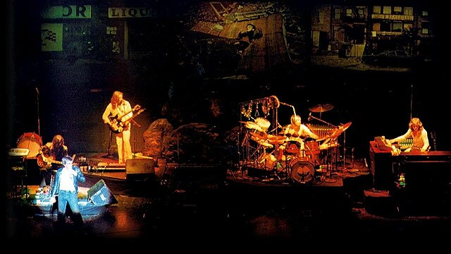 Genesis in their "classic lineup" incarnation on the Lamb tour in a wide shot in colour, 1974-75, clockwise from left: Peter Gabriel, Steve Hackett, Mike Rutherford, Phil Collins, and Tony Banks.
