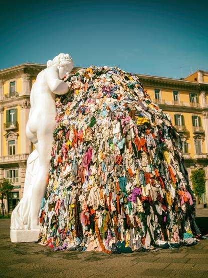 Eine Skulptur auf einer Plaza in Lissabon, die eine riesigen Berg an alten Kleidungsstücken zeigt. Daneben eine weiße , antike Figur.