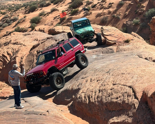 A red jeep on a steep angle being spotted by a man in front. Behind the red jeep is a seafoam green colored TJ.