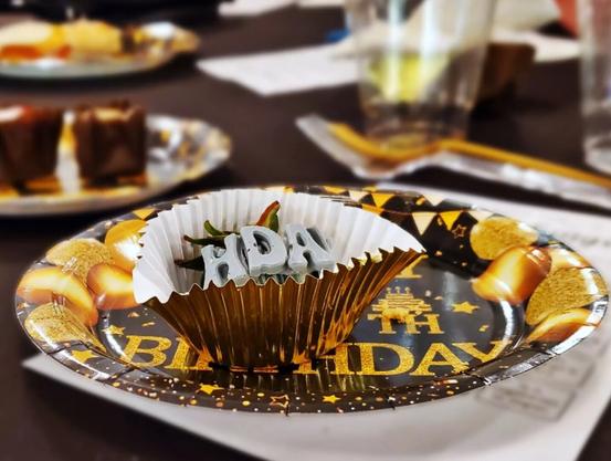 Photo of a black and gold paper plate with a small chocolate covered strawberry  with chocolate letters "H DAY" on top. The background is blurry but more desserts and cups can be seen.