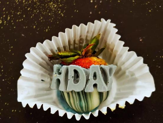 Detain of the chocolate covered strawberry, shot from above. The strawberry, resting in a pleated paper cup, has been dipped in white chocolate with blue stripes. The white chocolate letters "HDAY" are clearly in view.
