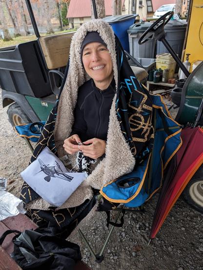 me sitting in a camp chair, bundled up in a fleece zipneck & hat and a hooded blanket, working on the scarf