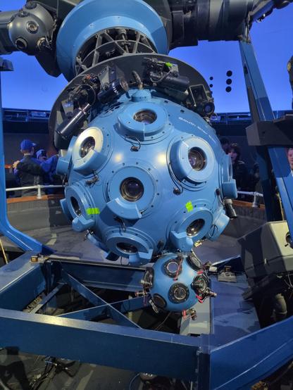 A large blue sphere with raised blue rings on the surface, each surrounding a glass lens. The sphere is mounted on a black metal truss.