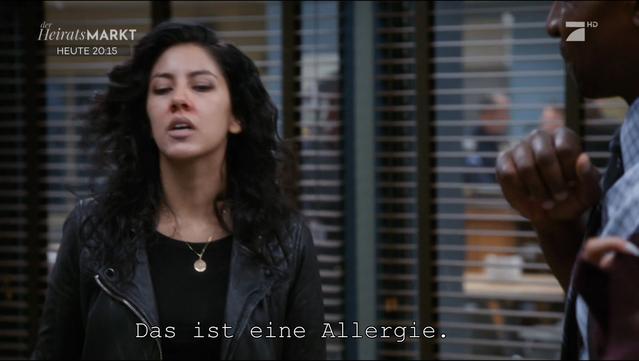 Description: The image shows a woman in a black jacket standing next to a man in a suit, both of them looking at each other. The woman is wearing a black dress and the man is holding an object in his hand. In the background, there is a window blind and at the bottom of the image there is some text which reads "Das ist eine Allergie".

Disclaimer: This image description was automatically generated and may not be entirely accurate.