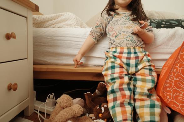 A child sitting on the edge of a bed, pointing down at some soft toys on the floor