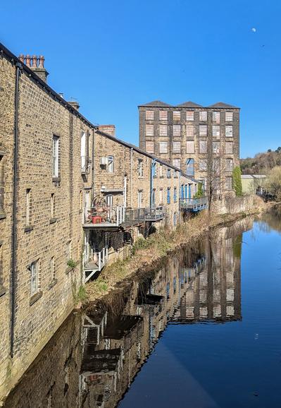 Stone built buildings backing directly onto a canal.  Buildings lead down towards an old textile mill.  Clear blue sky above with a bird flying "close" to a half moon.  All reflected, modulo small ripples, in the canal.