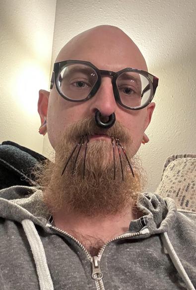 A bald individual with glasses features prominent facial piercings, including a septum ring and multiple metal ring-spike piercings in their lip. The person is wearing a gray zip-up hoodie and is seated in a well-lit indoor environment.