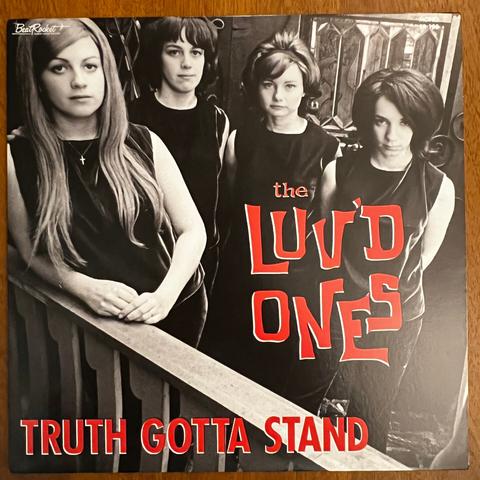 The Luv'd Ones front cover – a photograph of four young women in the 1960s, all wearing identical dark sleeveless tops. One stands to the left, a bit in front of the others, leaning on a wooden railing. Her hair is light colored, long, parted on one side, and she has a small cross on a necklace. The other three stand further back. The first has shorter, darker hair; the second has lighter hair, also shorter in a typical '60s cut with a headband, and the last has lots of dark hair in a '60s flip.