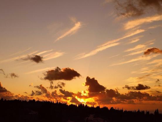 Ein warm orange-blauer Himmel während des Sonnenuntergangs. Die Sonne scheint noch durch einige Wolken am unteren Bildrand über dem dunklen Horizont. Dunkle und helle Wolken verteilen sich wie Pinselstriche über die Himmelsfläche.