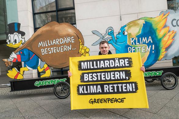 Ein Greenpeace Aktivist hält ein Banner mit der Aufschrift 'Milliardäre besteuern, Klima retten' vor einem Wagen auf dem Dagobert Duck mit Geldsack zu sehen ist und eine brennende Weltkugel versucht mit einer Schere den Sack zu zerschneiden.