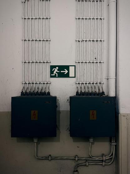 Two electrical junction boxes with multiple cables connected, mounted on a wall beneath an emergency exit sign pointing right.