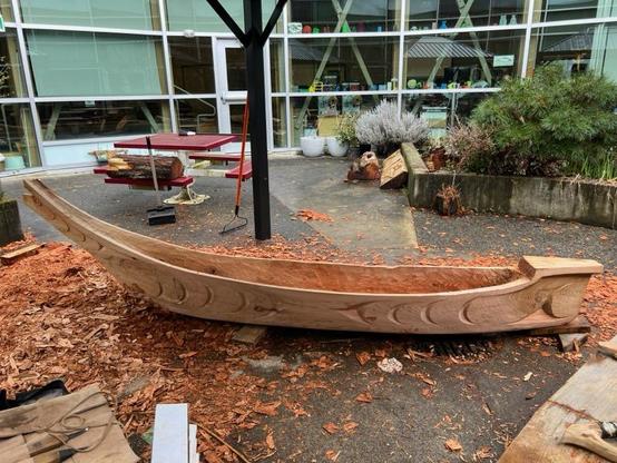 Medium shot of a carved, unfinished wooden dugout cedar canoe. The canoe is the primary focus, positioned horizontally in the foreground and running the length of the picture. The ground is splattered with wood shavings. The background includes a picnic table on the left with a log laid across it, plants, and a modern building with reflective windows. The lighting is overcast, with cool tones dominating the scene. The composition emphasizes the craftsmanship of the canoe against the backdrop of a contemporary school environment.