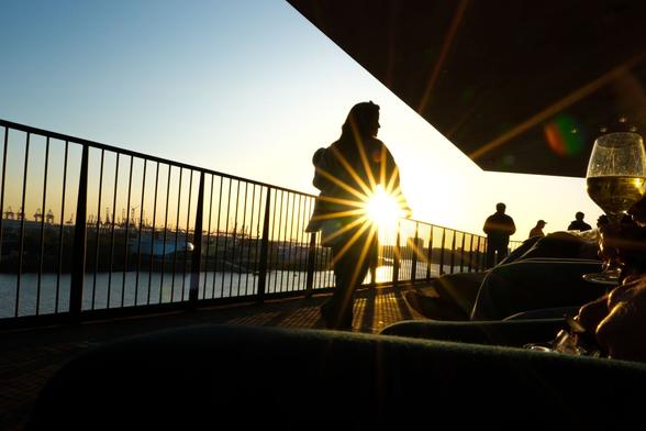 Kontrastreiche Aufnahme im Gegenlicht kurz vor Sonnenuntergang. Eine Frau läuft vor einem Geländer vom Betrachter weg und schaut dabei leicht nach rechts. Unter ihrem vorschwingenden Arm bildet die Sonne einen Blendenstern. Über ihr eine dunkle Decke, die den rechten oberen Bildteil einnimmt.An der rechten Bildseite sitzen Menschen, von denen man nur die Beine und Füße sieht, sowie ein einzelnes Glas Weißwein, das am rechten Bildrand mittig ins Bild gehalten wird und durch das an einer Stelle das Licht der Sonne scheint. Im Hintergrund Silhouetten weiterer Personen. Hinter dem Geländer eine Hafenlandschaft