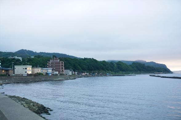 Ocean on a cloudy evening with a few buildings on the left and a hilly forest in the background.