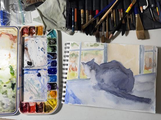 Photo: watercolor painting of a cat crouched on table with tail extending behind, looking out window.  although it's really a black cat his fur was seen as dark gray and light. To the left of the horizontal painting is the watercolor paintbox, vertical in the scene, with areas of color around the edge and two mixing areas in the middle. Above, an array of brushes in a black roll up holder point down at the painting.