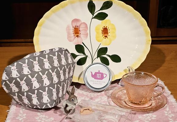 Tea tableau with a small bunny figurine positioned beside a miniscule pink princess style telephone as if waiting for it to ring. There are a pink Depression glass cup and saucer, a tea cozy patterned with smirking rabbits, and a large platter as a background that has one pink and one yellow handpainted flower on it. The tea is Vanilla Rose Chai from TCT.
