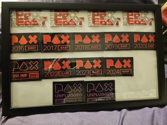 A framed collection of embroidered patches from PAX East and PAX Unplugged gaming conventions. The patches are neatly arranged in rows by year, spanning from PAX East 2012 to 2024, with a PAX Online 2021 patch and PAX Unplugged patches from 2022 and 2023 at the bottom.