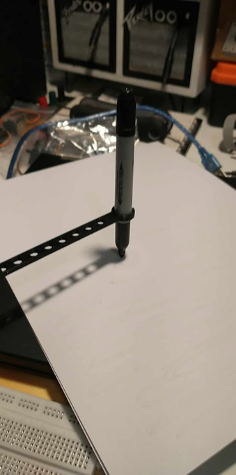 Image shows the actual arm as depicted in the computer aided drawing holding a Sharpie pen, the pen that was chosen as it gave good results. Arm holding pen shown with tip of pen sitting on a sheet of paper. I moved the arm manually back and forth to simulate the servo motor and was pleasantly surprised with the results.