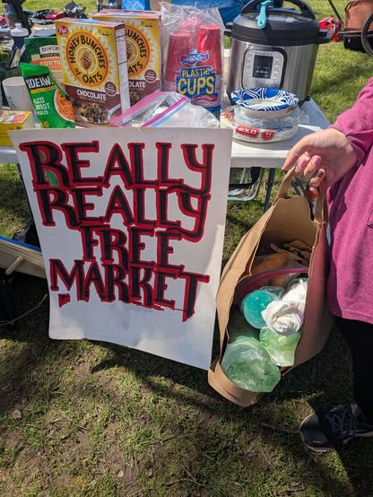 Collapsible white table with various food items, and a crockpot with a hanging sign that says "Really Really Free Market" and an erased word below.  Me standing next to it with a bag of hot drink cups, and croissants.