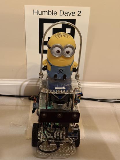 GoPiGo3 robot with Minion character Dave on top, on his dock in front of a sign with the robot's name "Humble Dave 2" and a large 6 by 6 square AprilTag.