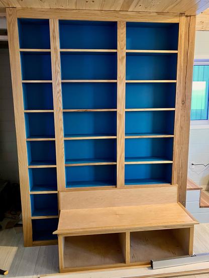 A set of built in shelves in a cabin. The face is red oak. The interior of the shelves is a deep blue. In front of the shelves is a built in bench.