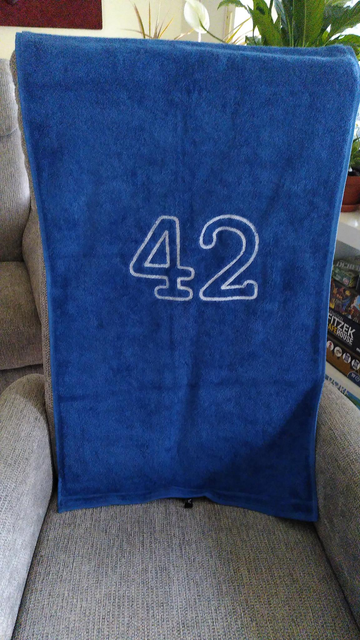 Ein blaues Handtuch, auf dem eine große "42" steht, hängt in einem Wohnzimmer-Kontext über der Lehne eines grauen Sessels.
Die 42 ist in der Mitte des Handtuchs aufgestickt, sodass die Ziffern aufrecht stehen, wenn das Handtuch hochformatig betrachtet wird. Die ungefähr 20 cm großen Zahlen sind als weiße Umrisse dargestellt.