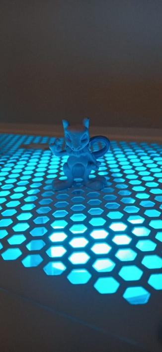 Foto de uma miniatura do pokemon Mewtwo em cima de uma estrutura de metal com diversos furos em formato circular, de onde sai uma luz azul claro, que o ilumina.