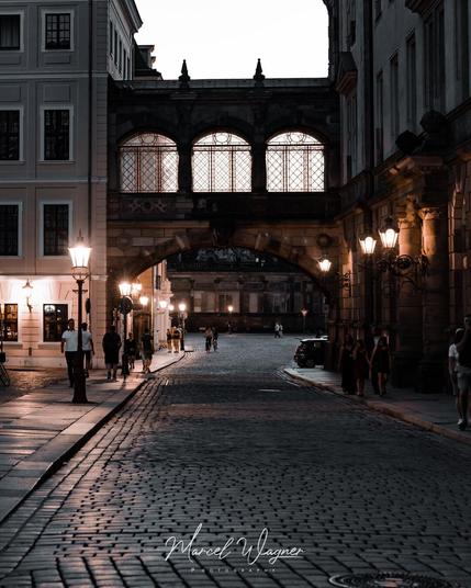 A dimly lit European street at dusk with cobblestone pavement, glowing lanterns, and people walking under a large stone archway with tall, illuminated windows above.