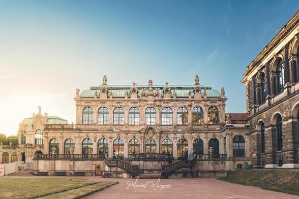 A symmetrical Baroque palace with large arched windows and ornate sculptures, lit by warm evening sunlight under a clear blue sky. The building is the Zwinger in Dresden, Germany.