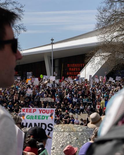 A far-off sign reading "Diversity Makes America Great", surrounded by protesters, with out of focus attendees in the foreground to the left and right.
