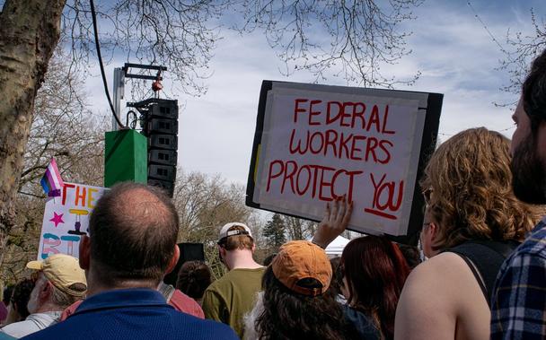 A woman holds a sign reading "Federal workers protect you" among a crowd of protesters at the "Hands Off!" rally.