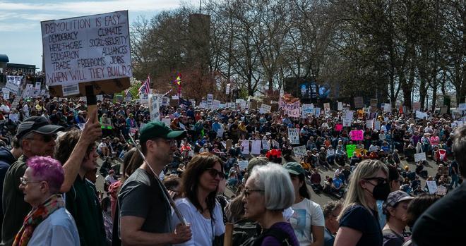 Photo of the throng of protesters in and around the International Fountain in the Seattle Center. A man to left holds a sign reading "Demolition is Easy - When will they build?" with various departments, civil and humans rights issues, and the word "democracy" all crossed out.
