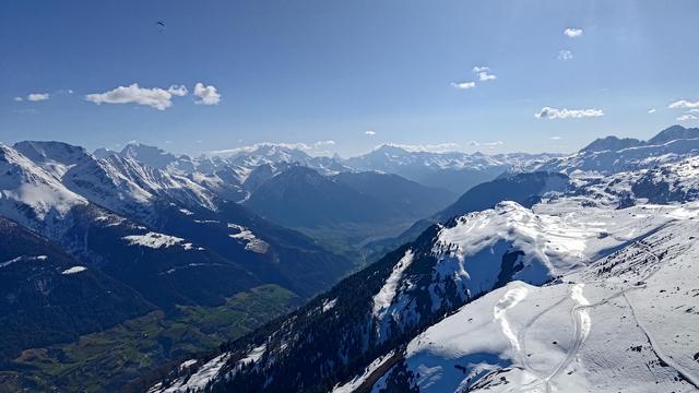 This is a stunning aerial photo taken from a paraglider high above Fiescheralp in the Swiss Alps. The foreground shows snow-covered ridges and ski tracks typical of the Aletsch Arena region, with steep slopes dropping down toward the green Rhone valley below. On the left, the dramatic peaks of the Valais Alps rise sharply, many still coated in spring snow. In the distance, you can spot iconic giants like the Matterhorn and Weisshorn, peeking through the layered ridgelines. A lone paraglider floats gracefully against the deep blue sky, perfectly capturing the serenity and scale of this alpine panorama.