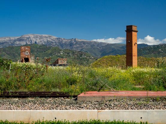 A brick chimney and ruins of other brick buildings against the background of a mountain range under a clear blue sky. They are surrounded by wild flowers and tall grass. Close to the camera are tram tracks.