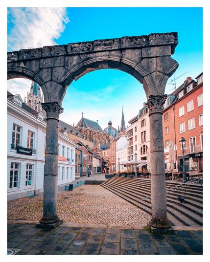 An urban street scene with the prominent Roman remains of a portico and the famous Aachen Cathedral in the background, showing the city's architecture and lively surroundings.