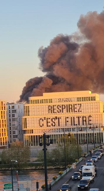 sur la façade du bâtiment au premier-plan, en grosses lettres visibles de loin «Respirez c'est filtré»... et en arrière-plan le ciel est noirci par la fumée épaisse d'un incendie d'un tout autre bâtiment