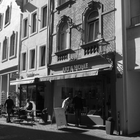Schwarzweißfoto. Zwei Altbau-Geschäftshäuser mit Schaufenstern im Erdgeschoss und Fensterreihen darüber. Vor dem Schaufenster im rechten Haus steht ein Paar und betrachtet die Auslagen. Vor dem Schaufenster im rechten Haus stehen Stühle und Tische. An einem sitzt ein Mann, ein anderer geht vorbei. Über dem rechten Schaufenster hängt eine Leuchtreklame "ART & HOME".