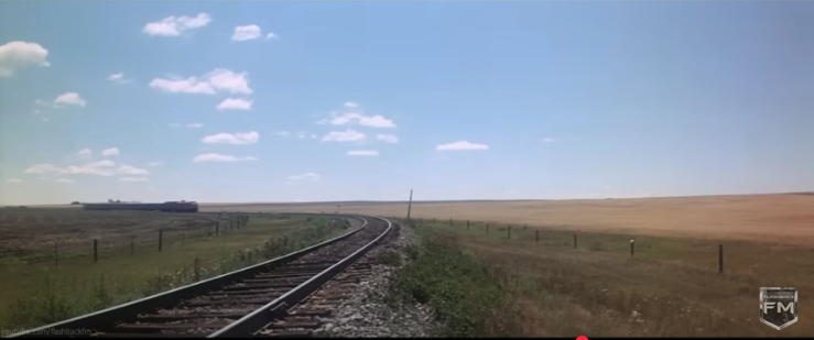 Screenshot of Youtube video showing a railroad track curving to the left in the distance and running across a yellowing prairie. A diesel train is approaching from afar and partially silhouetted against a blue sky with a scattering of white cotton-wool clouds.
