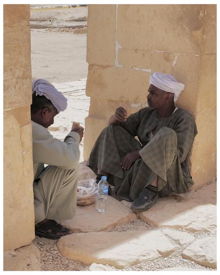The image shows two men engaged in a conversation. They are sitting on the ground in the corner of a structure made of yellowish stone or bricks, possibly in a desert or arid area given the sandy and barren landscape visible in the background. Both men are wearing traditional attire, which includes turbans and long robes, indicating that the setting could be in a region where such clothing is customary, perhaps somewhere in the Middle East or North Africa. A water bottle is also visible on the ground near them, and both men are holding pieces of Egyptian bread.  The overall scene suggests a casual, perhaps spontaneous, gathering in a tranquil environment.