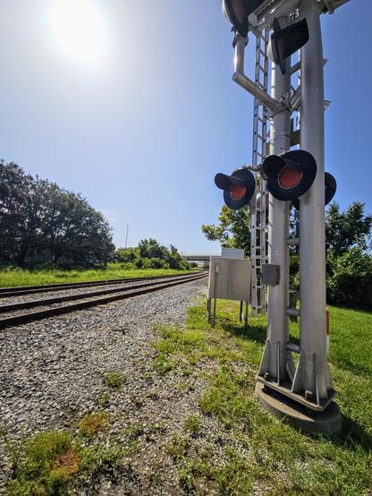 Beneath a glaring, burning hot mid day sun, a view down an old rusty set of railroad tracks on a bed of grey gravel next to a tall crossing alert lights and arm.