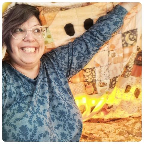 en premier plan je vous souris, en chemise de nuit, avec mes lunettes, et je lève le bras pour soulever la couverture patchwork et vous inviter dans la cabane magique !
